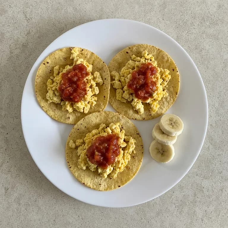 Three folded corn tortilla breakfast tacos with scrambled eggs and salsa on a white plate, with sliced banana on the side.
