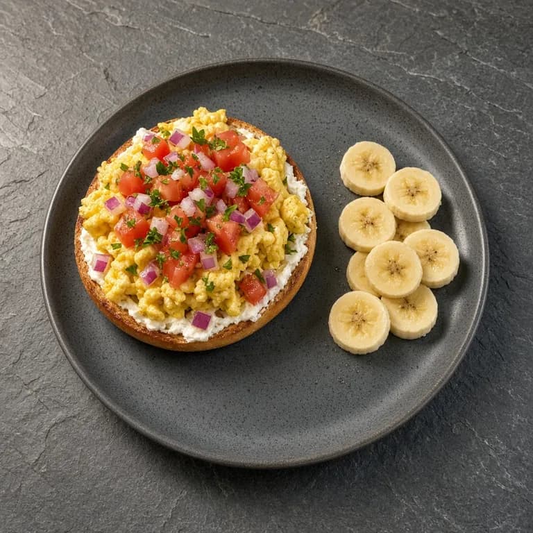 Toasted bagel halves with cream cheese, scrambled eggs, tomato and red onion on a white plate, with sliced banana on the side.