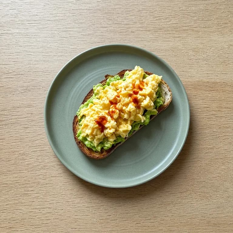 Two slices of sourdough toast spread with mashed avocado, topped with scrambled eggs and drizzled with red hot sauce, on a white plate.