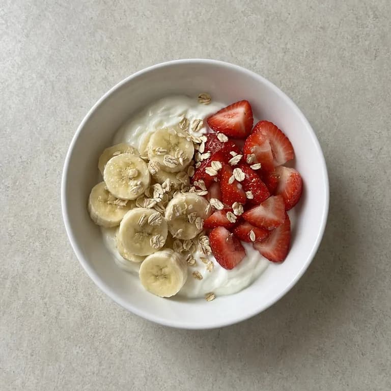 White bowl of Greek yogurt with strawberries, sliced banana and dry rolled oats on top.